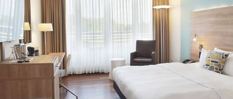 Et hotelværelse med seng, skrivebord med computer, stol, lampe og et stort vindue med gardiner.
