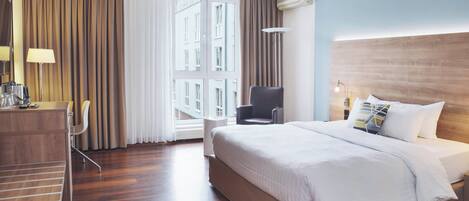 A hotel room with a large bed, a desk with a chair, a small table, and a window with curtains.
