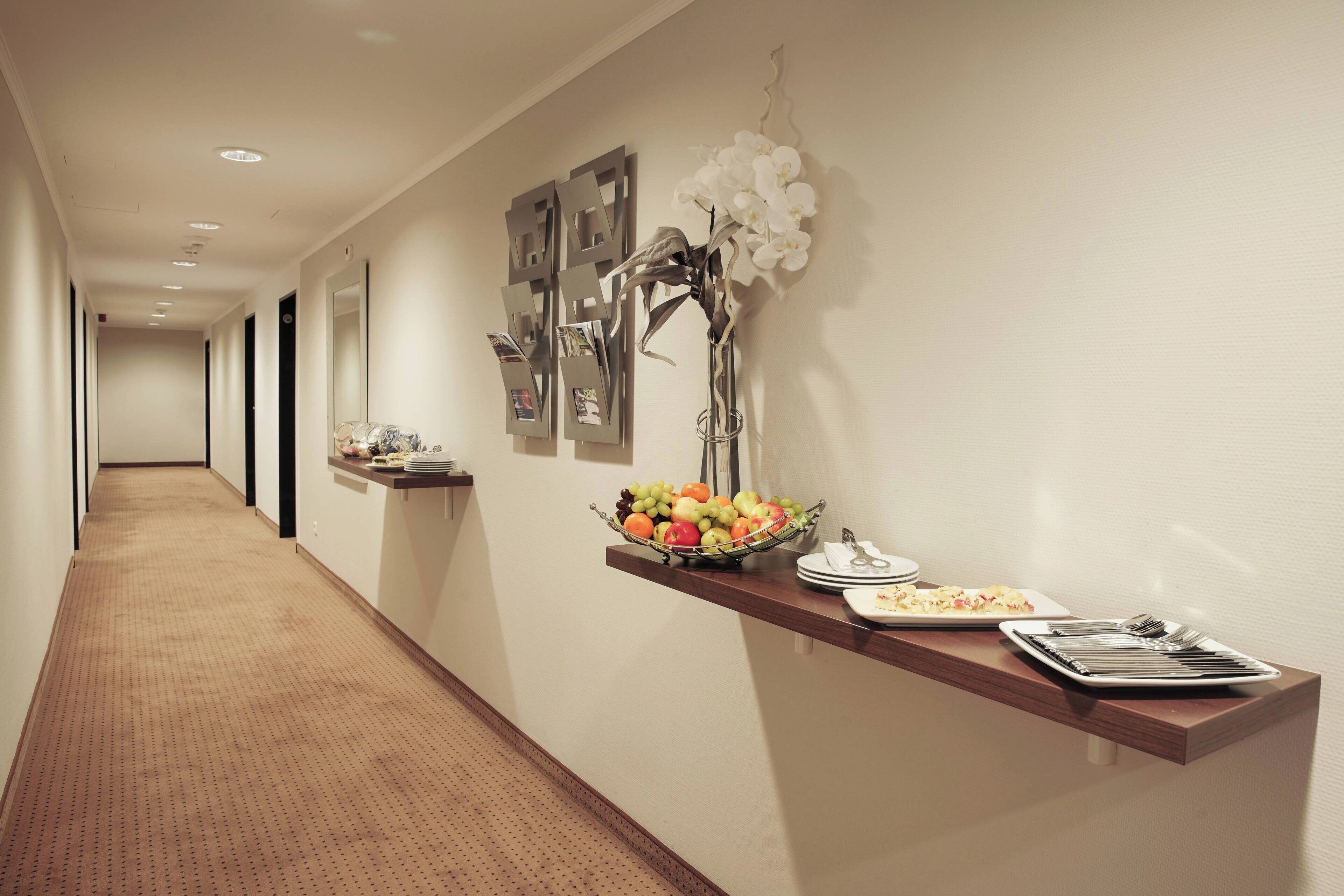A hotel corridor with a reception desk displaying fruits, a vase of flowers, and framed pictures.