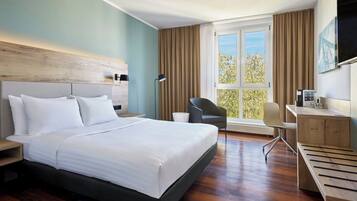 A modern hotel room with a large bed, a desk, a chair, and a window with curtains.