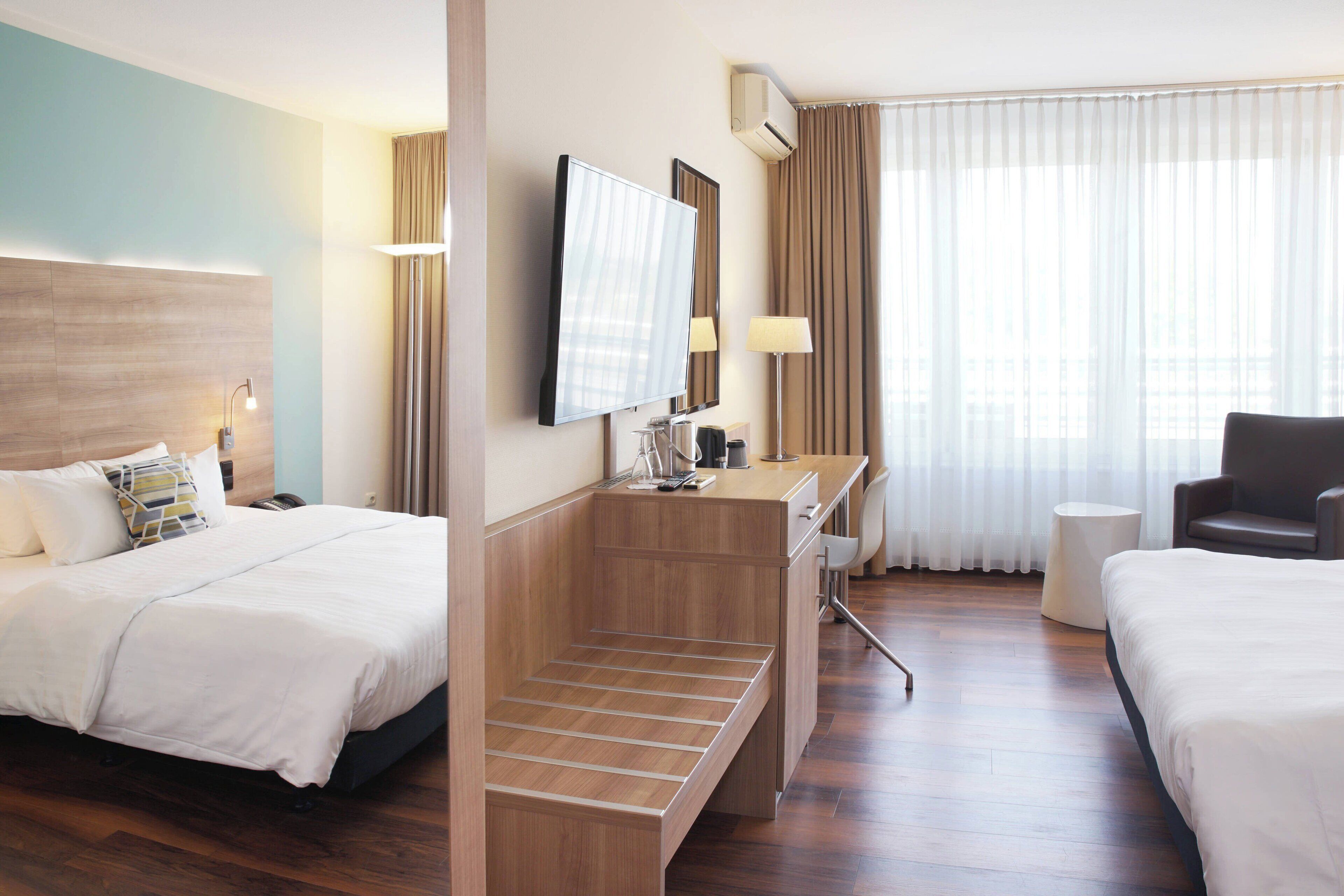 Una habitación de hotel con dos camas, un escritorio, una silla, un televisor y un ventanal con cortinas.