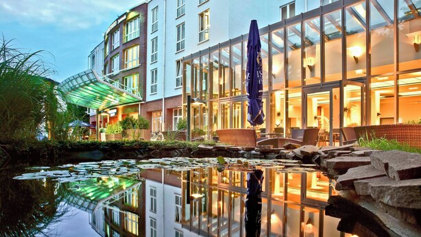 A hotel with a glass facade, outdoor seating, and a pond with lily pads. - Courtyard by Marriott Dresden (Dresden)