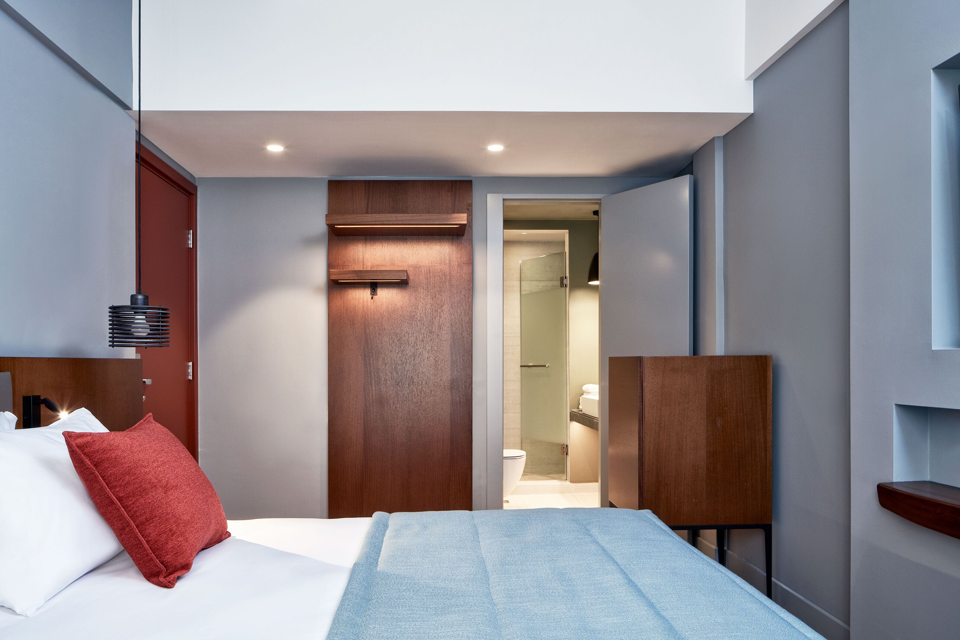A modern hotel room with a bed, a red pillow, a wooden headboard, a nightstand, and a built-in shelf.