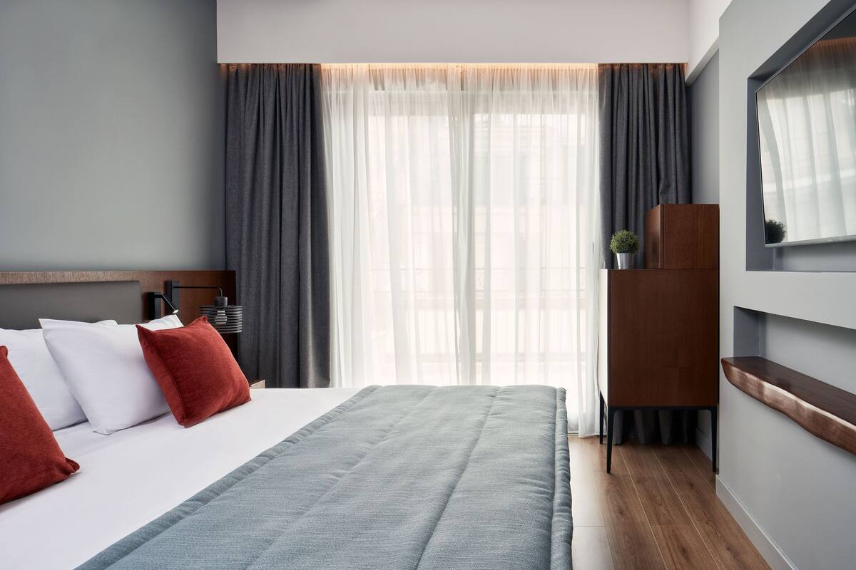 Moderni hotellihuone, jossa on suuri sänky, harmaat verhot, puinen yöpöytä ja pieni ruukkukasvi.