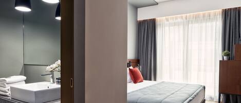 A modern hotel room with a large bed, a bedside table, a mirror, a bathroom with a sink and towel rack.