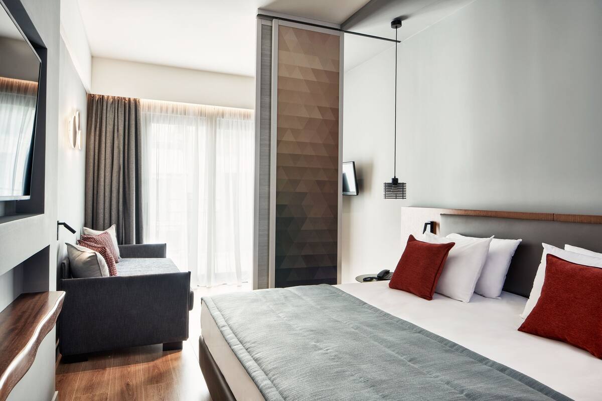 Moderni hotellihuone, jossa on sänky, sohva, televisio ja suuri ikkuna, jossa on verhot.