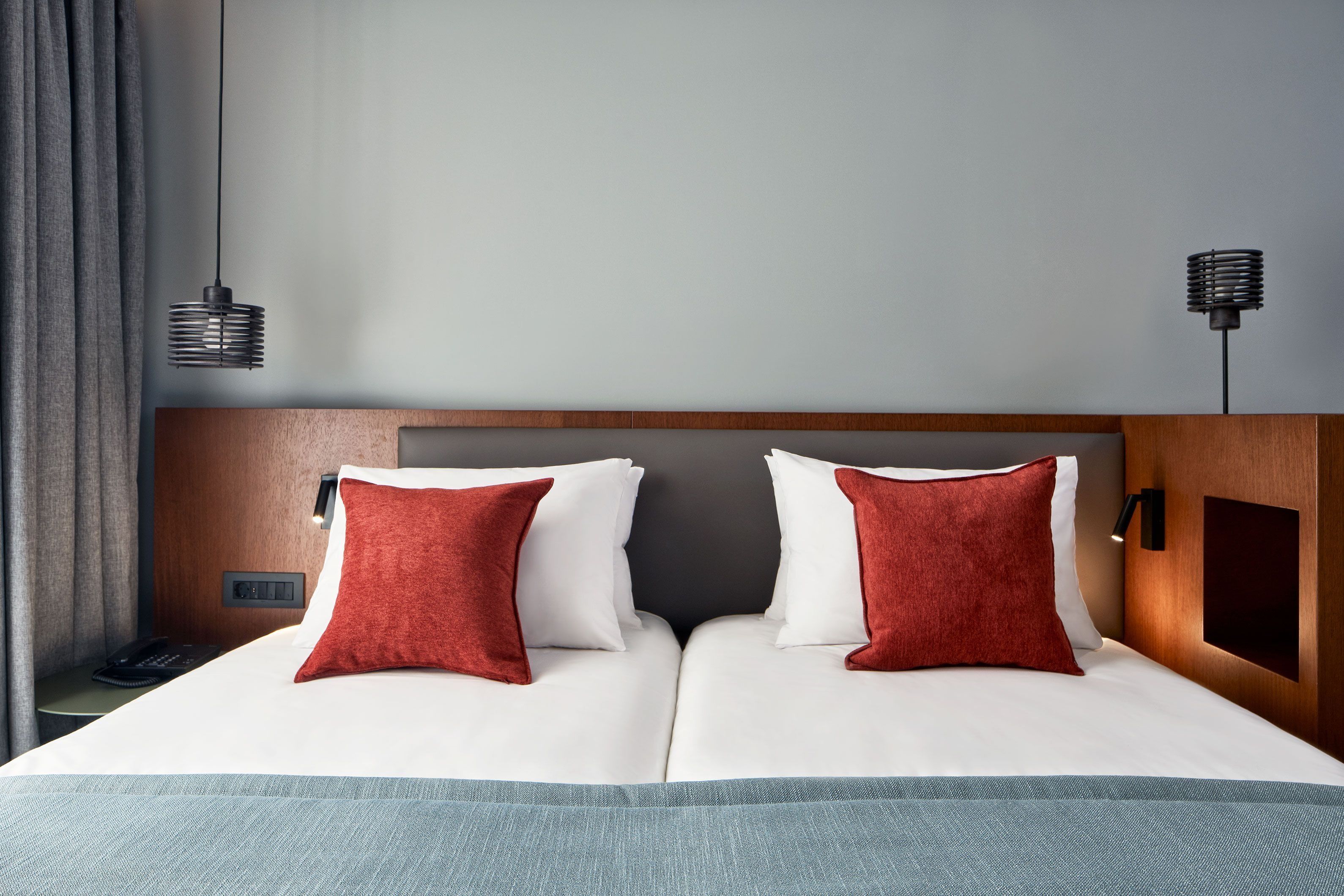 A hotel room with a neatly made bed, two red pillows, a wooden headboard, a bedside table with a phone, and a wall-mounted lamp.