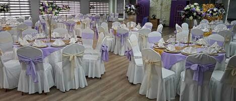Un salón de banquetes con mesas redondas, sillas blancas con cintas moradas y centros de mesa florales.