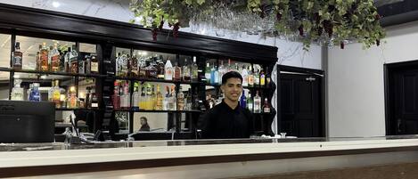 Un espace bar avec un comptoir, des étagères remplies de bouteilles d’alcool et une plante suspendue ornée de raisins.