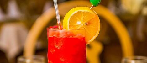 Un grand verre rempli d’une boisson rouge, avec une paille et une cerise enfilée sur une brochette, le tout décoré d’une tranche d’orange.