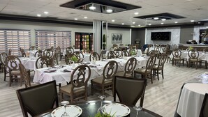 A spacious dining area with multiple tables and chairs, a central column, and a television screen on the wall. - La Quinta Hotel (La Ceiba)