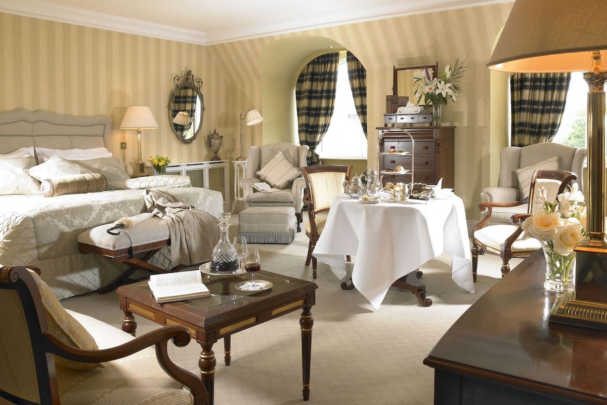 A classic, elegantly furnished bedroom with a large bed, a sitting area with a table, and a dining area with a three-tiered stand.