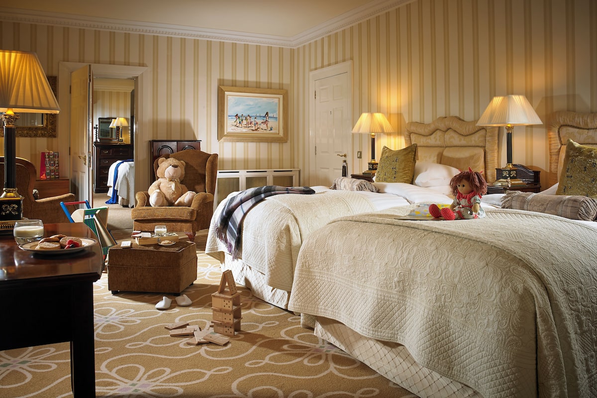A hotel room with a large bed, a sitting area with a chair and a table, a dresser, a mirror, and a painting on the wall.