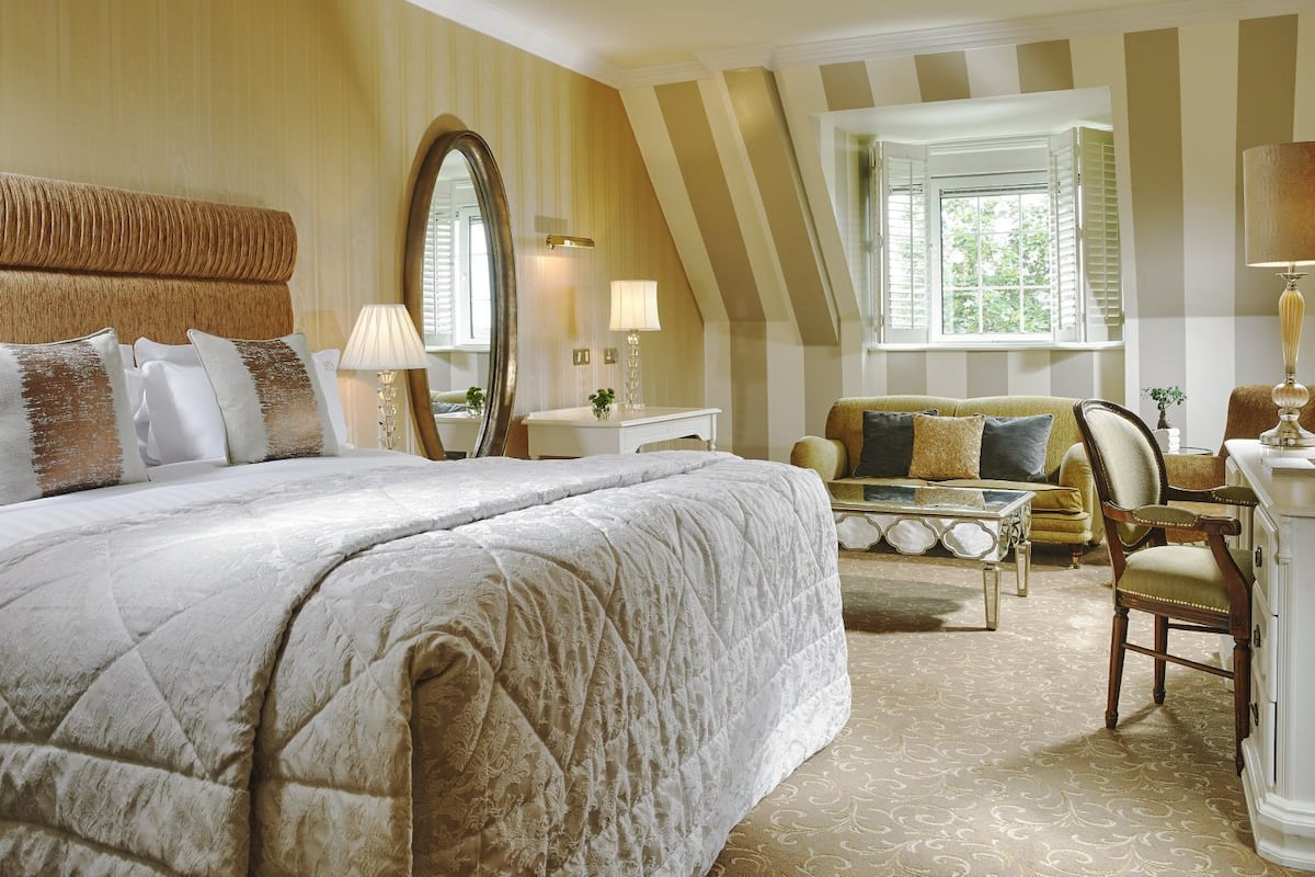 A bedroom with a large bed, a sitting area with a sofa and armchairs, a mirror, and a window with shutters.