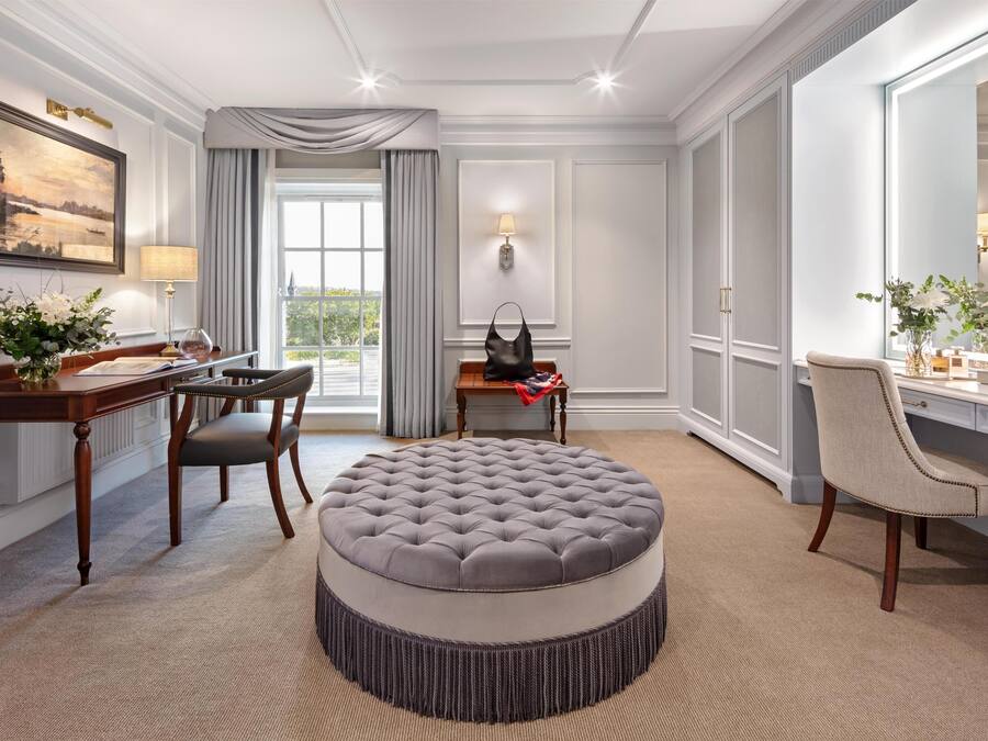 Une chambre d’hôtel avec un pouf rond, un bureau avec une chaise, un grand miroir et une vue sur l’extérieur.