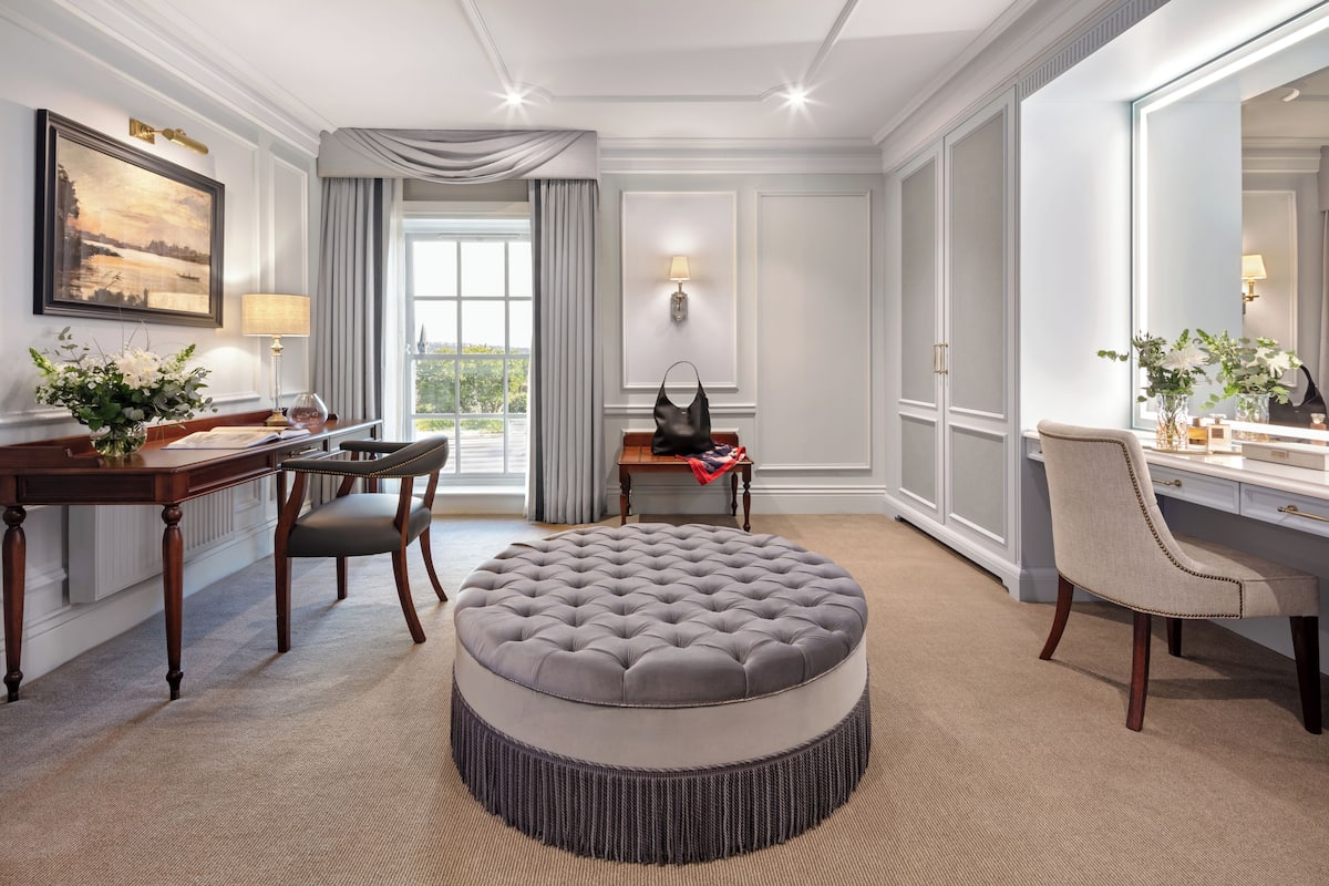 A hotel room with a round ottoman, a desk with a chair, a large mirror, and a view of the outdoors.