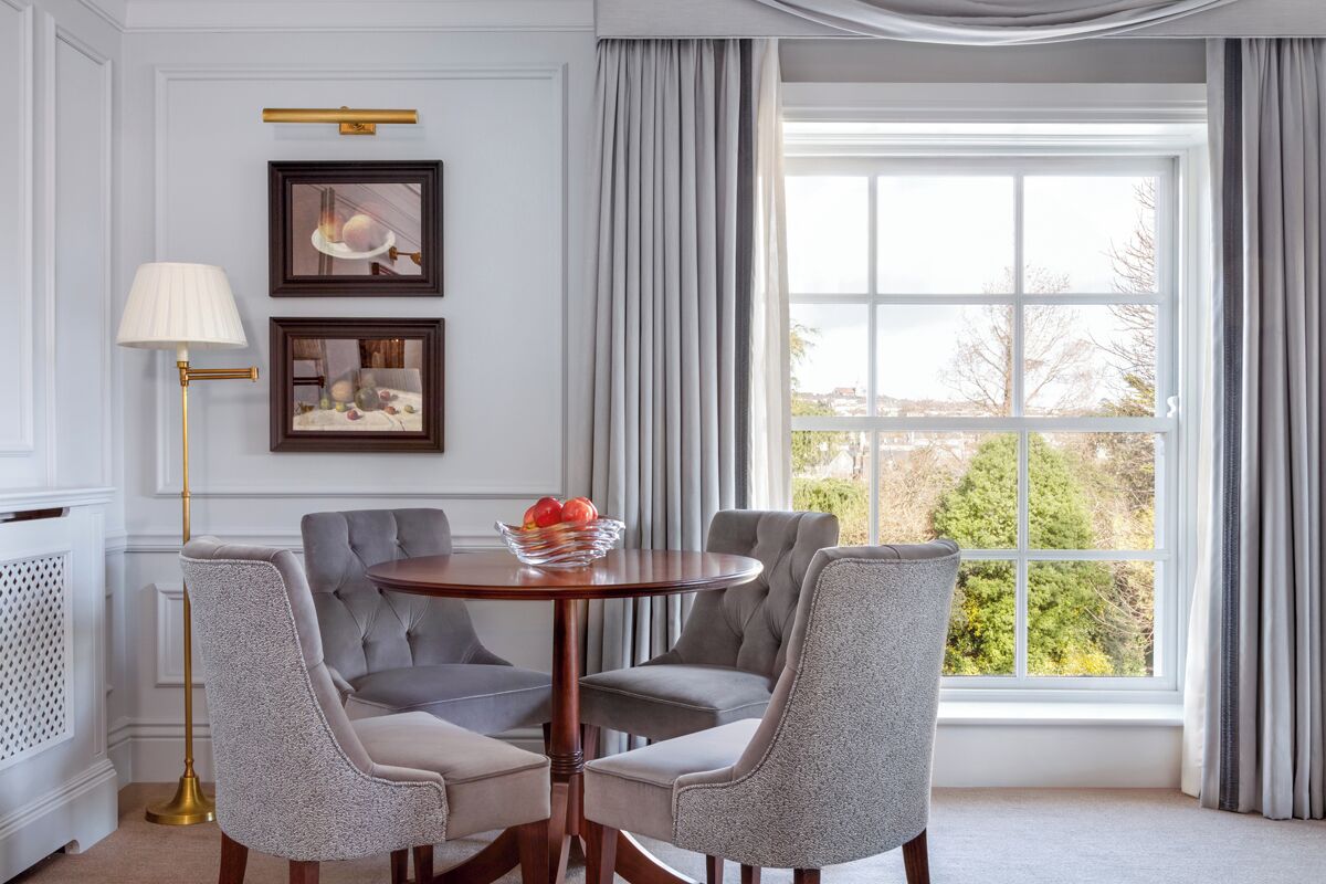 A dining area with a round table, four grey upholstered chairs, a floor lamp, and two framed pictures on the wall.