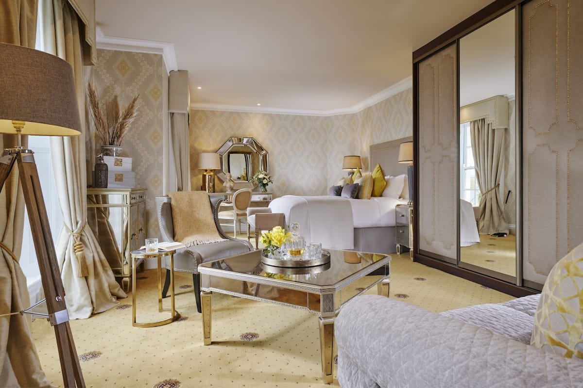 A luxurious hotel room with a large bed, a sitting area with a sofa and armchairs, a dining table, and a large mirror.