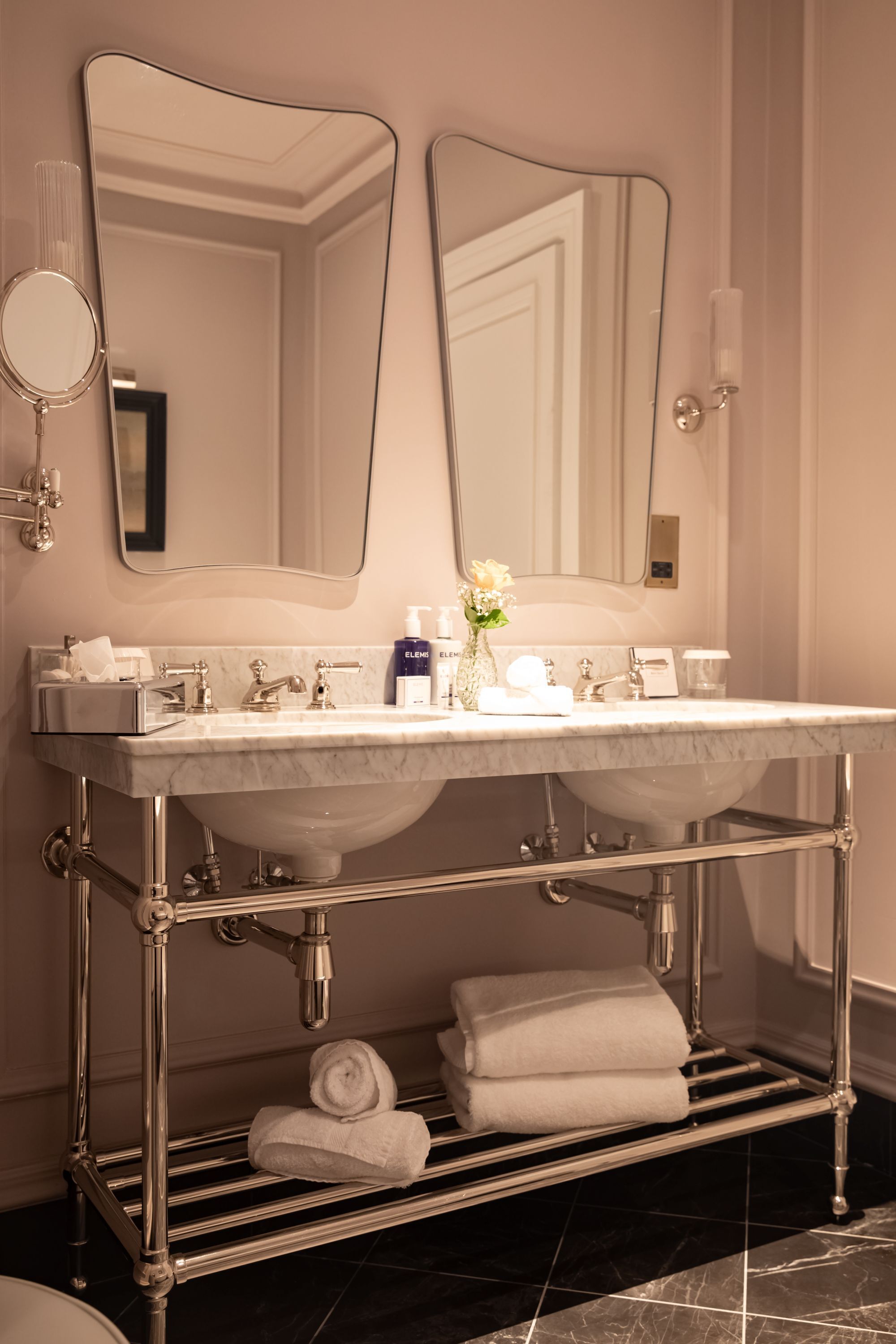 A bathroom with a double sink, two mirrors, a towel rack, and a vase with flowers.