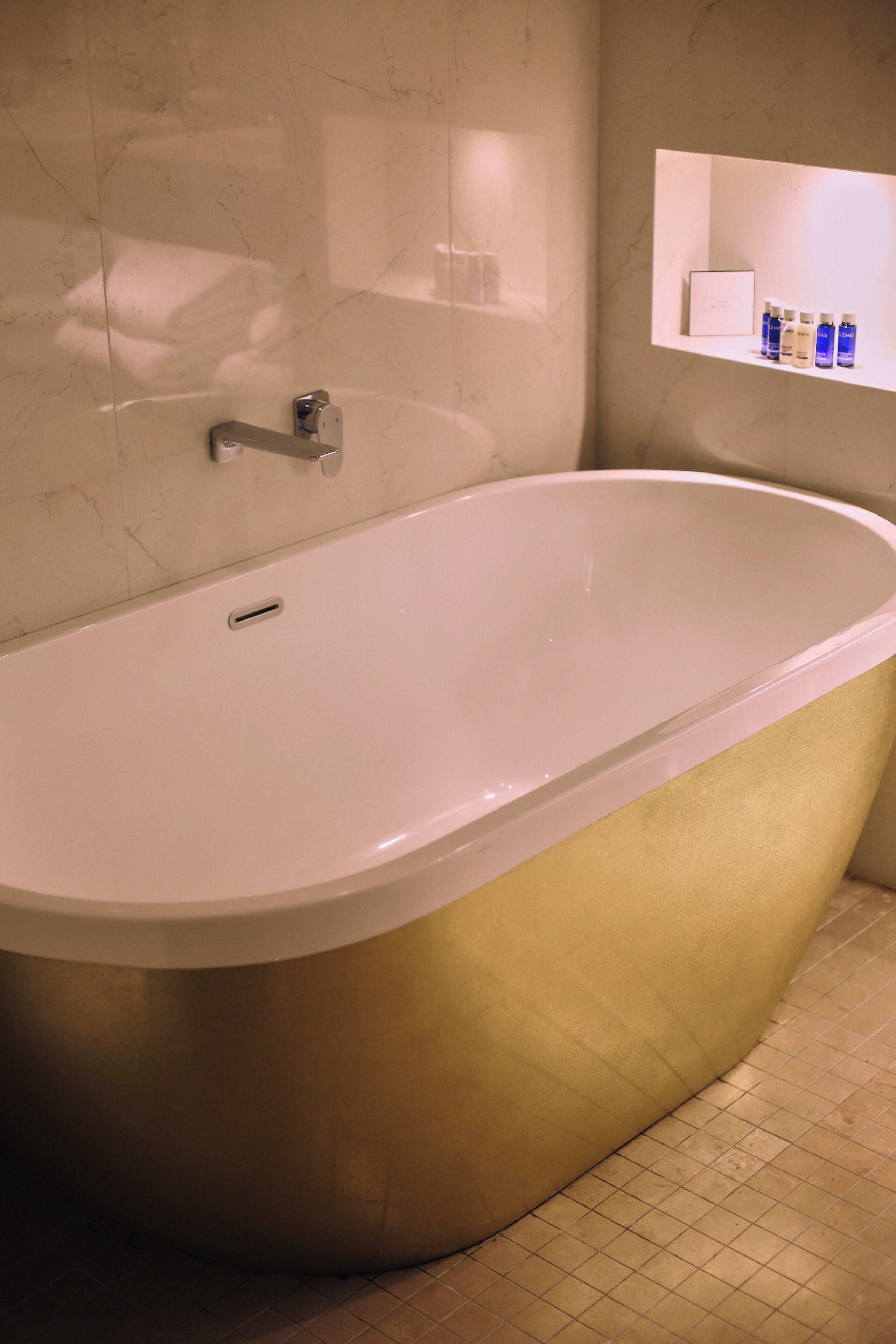 A modern bathroom with a freestanding bathtub, a wall-mounted shelf with toiletries, and a tiled floor.