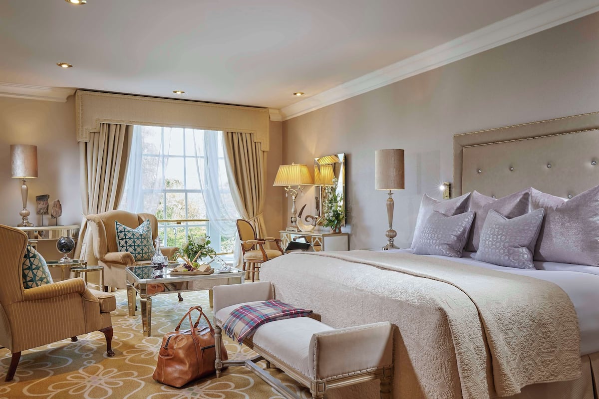 A well-appointed hotel room with a large bed, a sitting area with a table and chairs, and a view of the outdoors through the window.