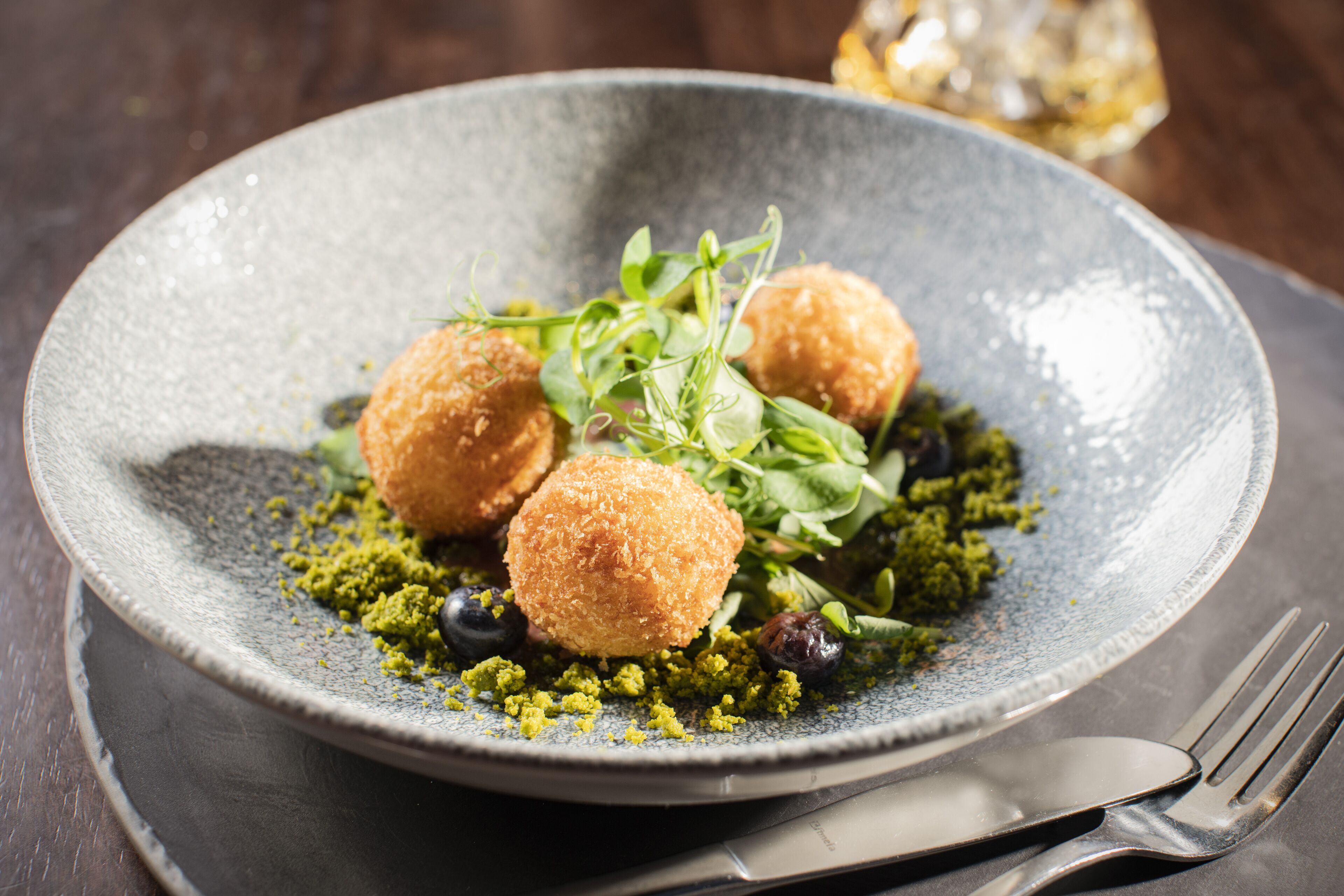 A plate with three round, golden-brown fried balls, green leafy garnish, and a scattering of green crushed nuts.