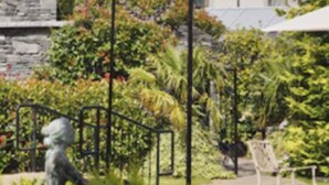 Un edificio que recuerda a un castillo, con base de piedra y estructura superior blanca, rodeado por un jardín bien cuidado con diversas plantas y una estatua.