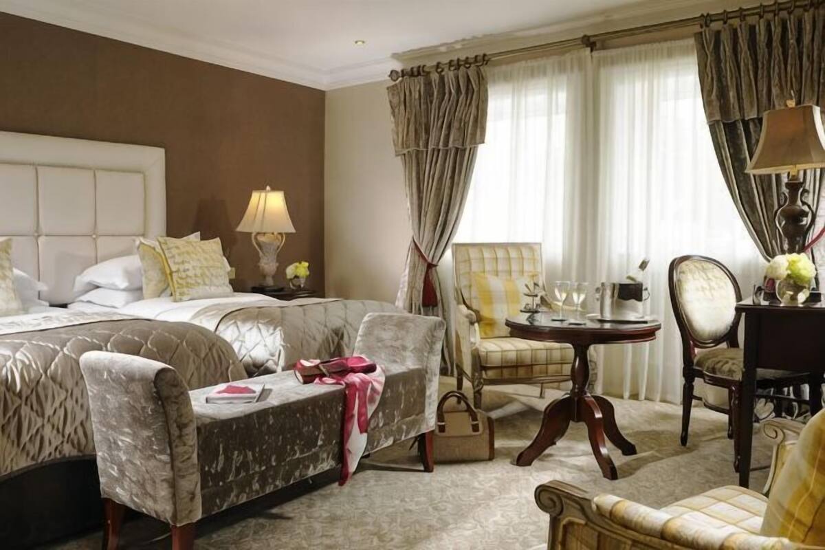 Habitación de hotel con una cama grande, dos sillones, una mesita y una mesita de noche con una lámpara.