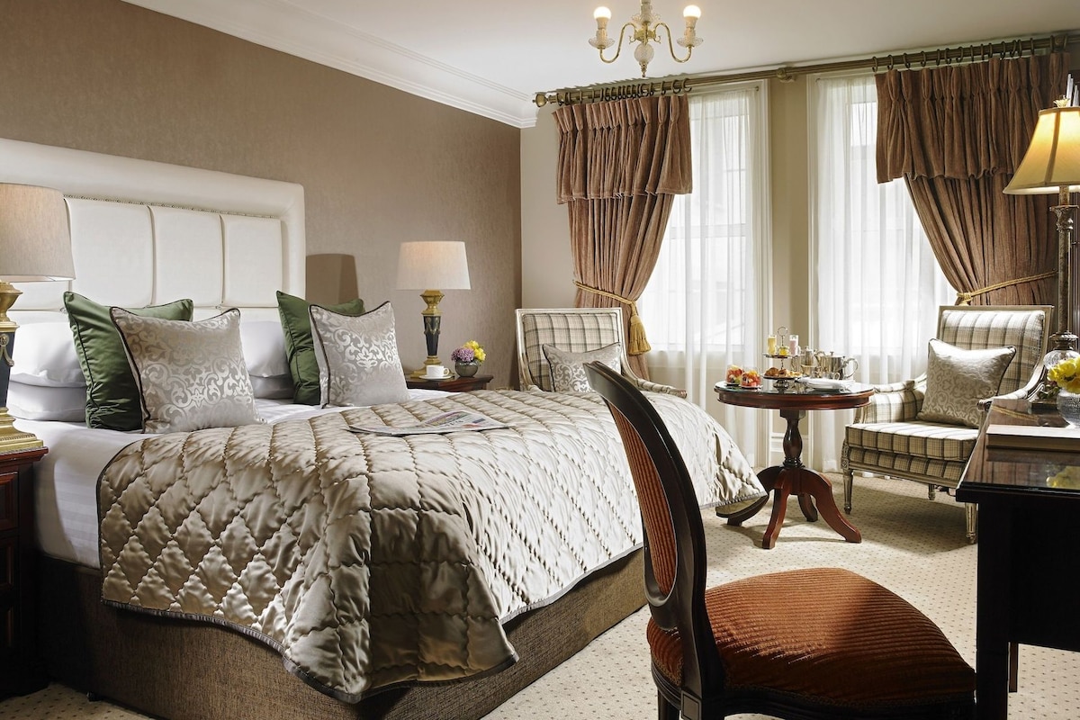 A hotel room with a large bed, two chairs, a small table with a lamp, and a chandelier.