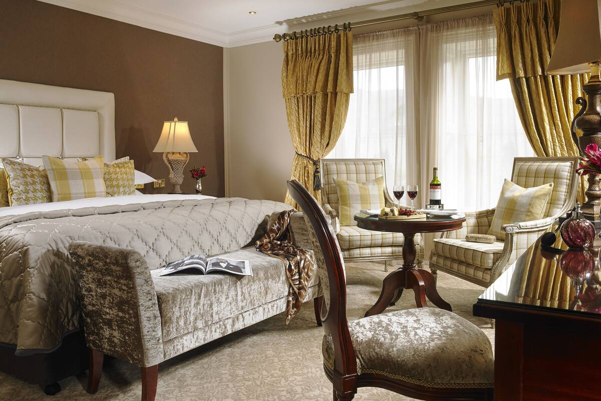 Habitación de hotel con una cama grande, dos sillones, una mesita con una botella y vasos, y una lámpara.
