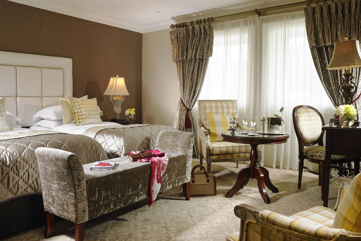 Una habitación de hotel con una cama grande, dos sillones, una mesita de noche con una lámpara y una silla con un florero.