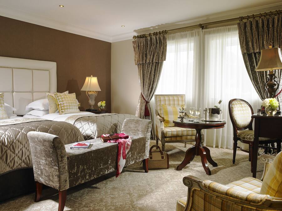 A hotel room with a large bed, two armchairs, a small table with a lamp, and a chair with a vase of flowers.