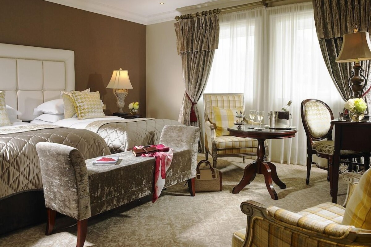 A hotel room with a bed, two armchairs, a small table with a lamp, and a window with curtains.