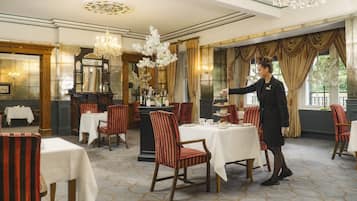 A formal dining room with tables set for service, a waiter in uniform, and classic decor including chandeliers and curtains.