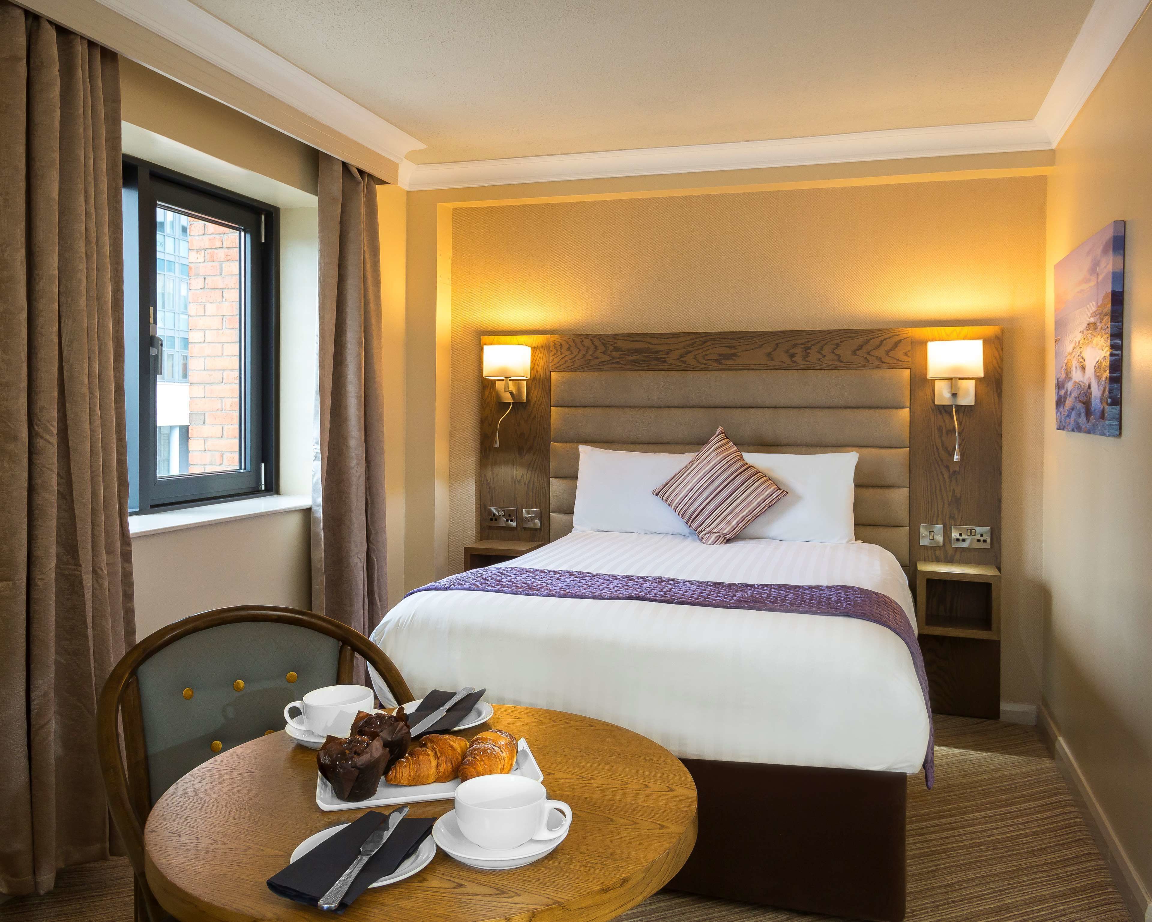 A hotel room with a bed, bedside lamps, a window with curtains, a dining table with pastries and a cup, and a painting on the wall.