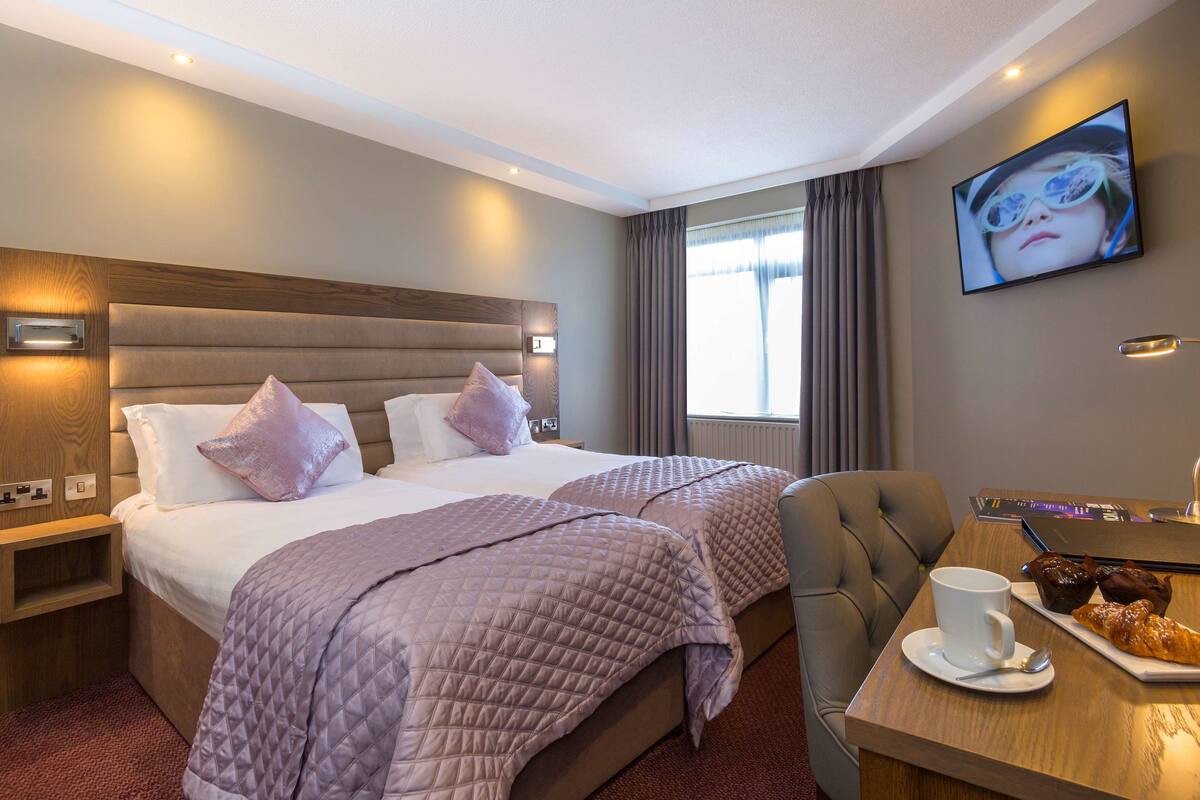 A hotel room with two beds, a desk with a laptop, a cup of coffee, and a croissant.