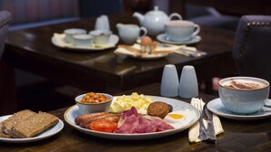 A breakfast setting with plates of eggs, bacon, tomatoes, and toast, along with a cup of coffee and a teapot on a table.