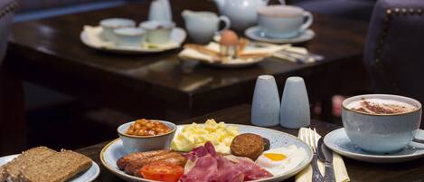 Un desayuno con platos de huevos, tocino, tomates y tostadas, junto con una taza de café y una tetera sobre una mesa.