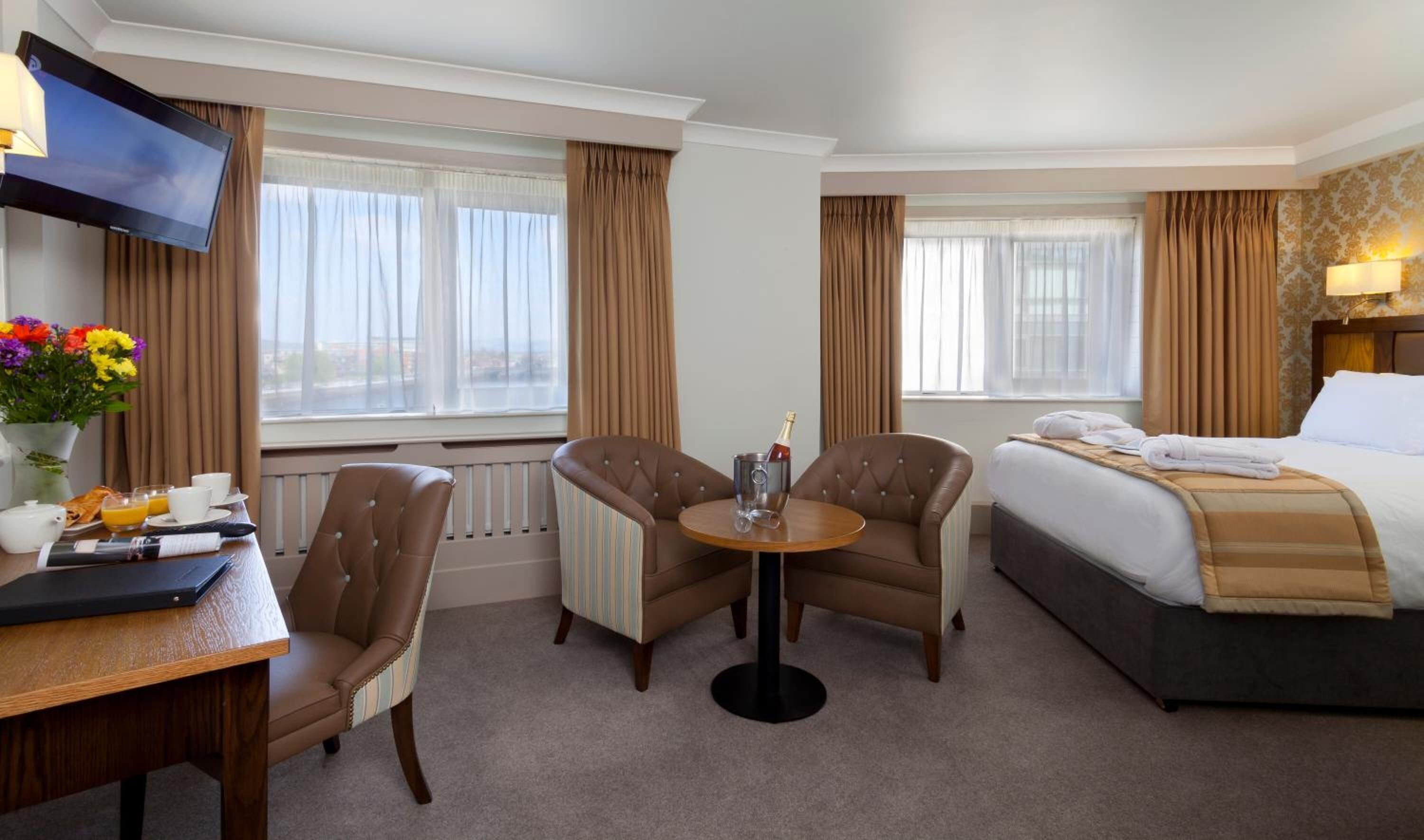 Ein Hotelzimmer mit einem Bett, zwei Sesseln, einem kleinen Tisch mit einer Champagnerflasche, einem Fernseher und einem Fenster mit Vorhängen.