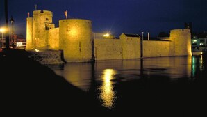 A historic stone fortress with multiple towers and walls, illuminated by street lamps, reflecting on the water.