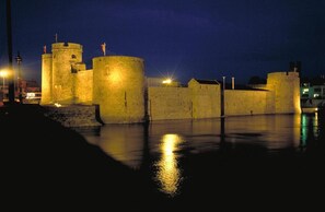 A historic stone fortress with multiple towers and walls, illuminated by street lamps, reflecting on the water.