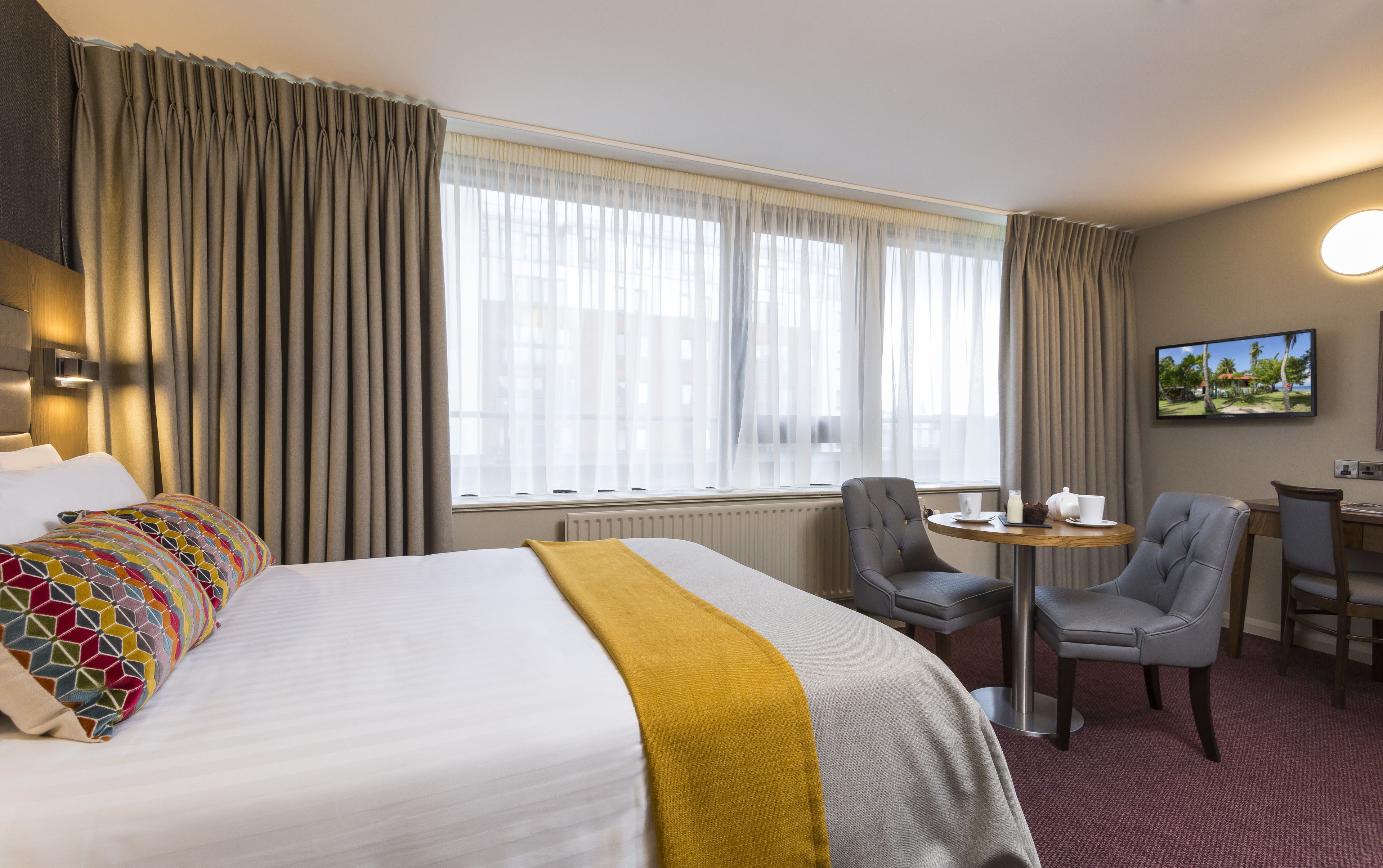 A hotel room with a large bed, a desk, two chairs, a television, and a window with curtains.