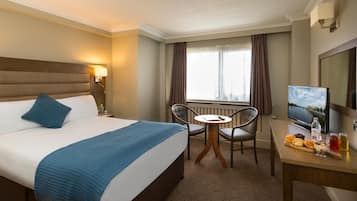 Una camera d'albergo con un letto, una scrivania con televisore, una sedia e una finestra con tende.