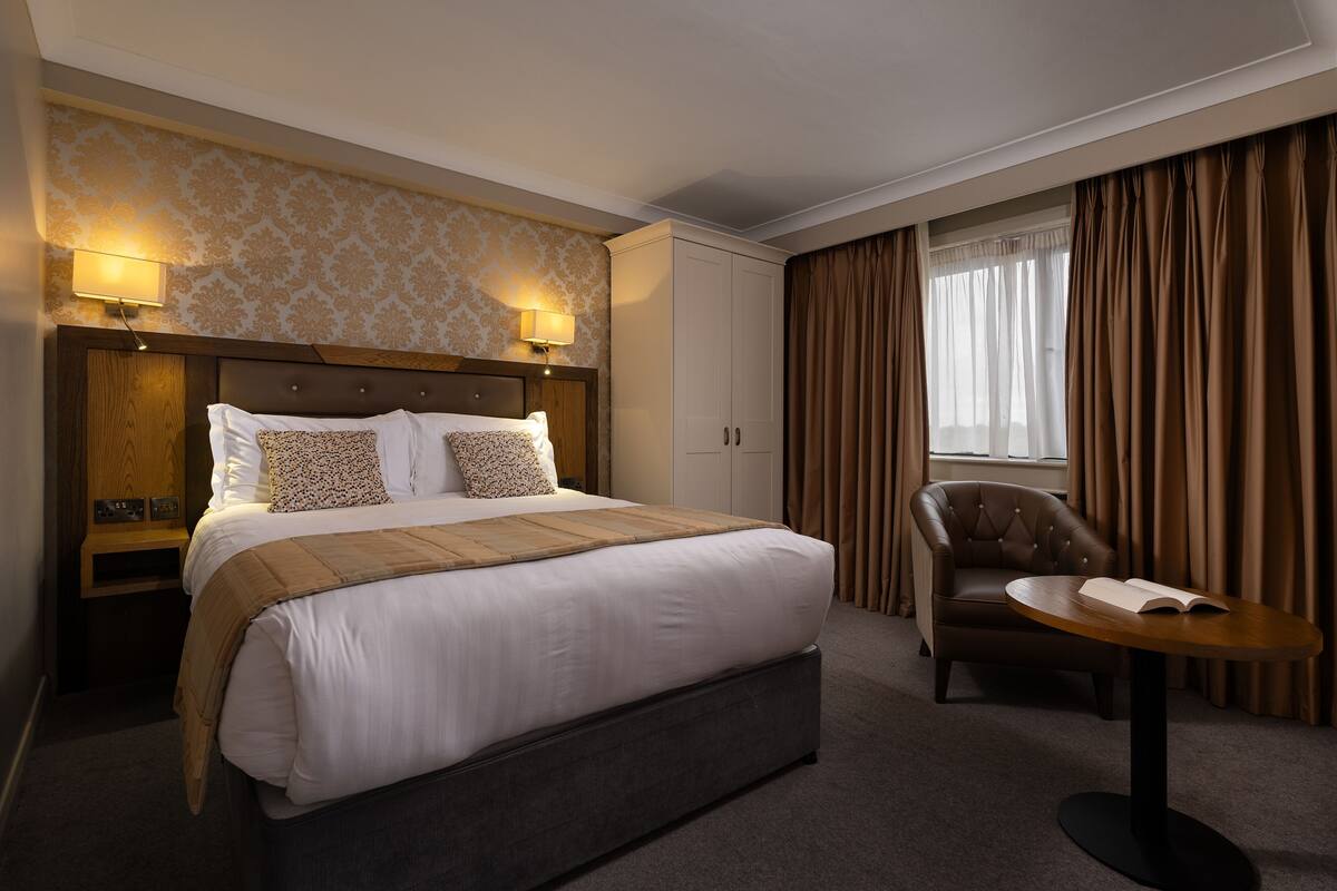 A hotel room with a large bed, a chair, a small table, and a lamp.