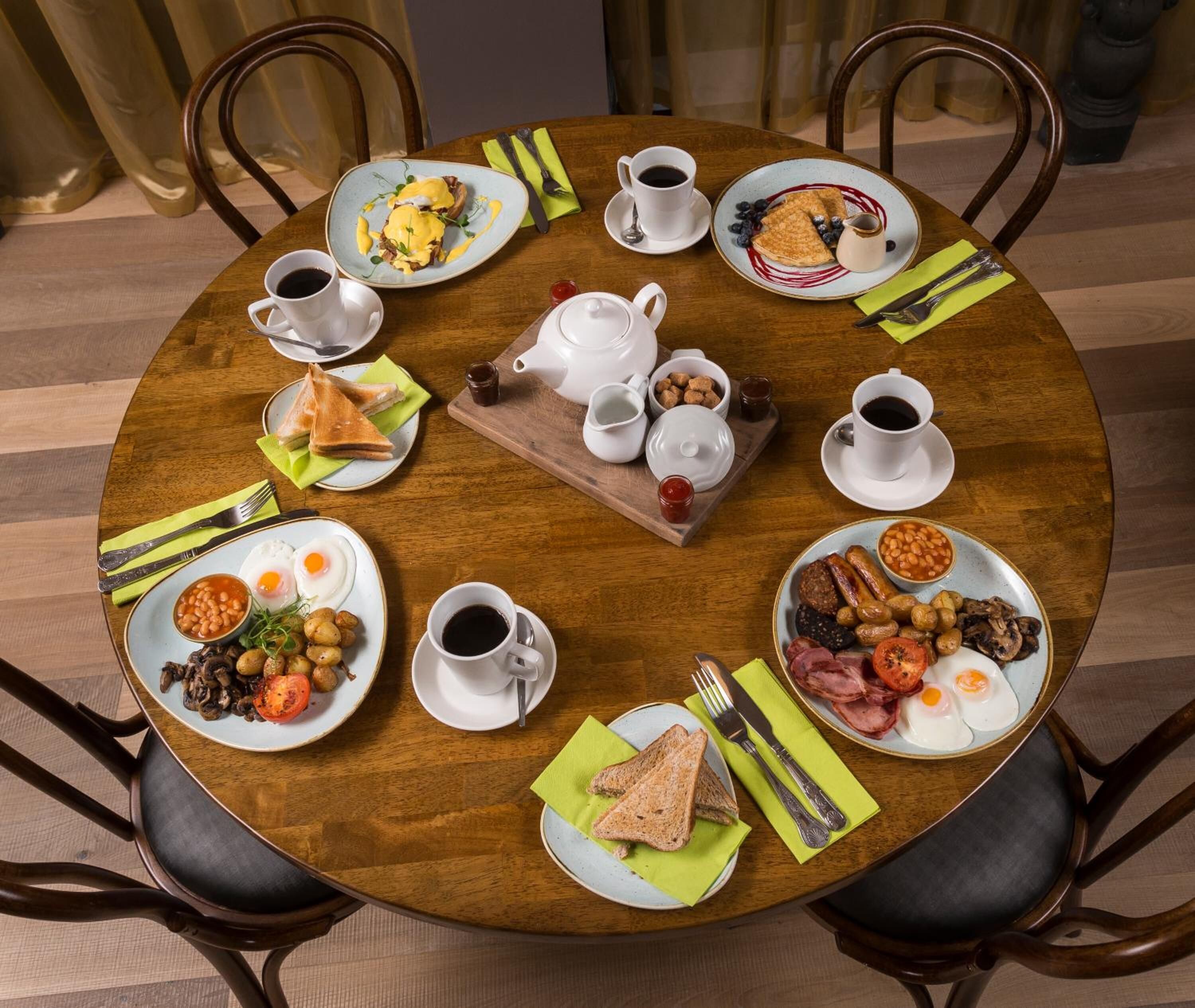 A round wooden table set for breakfast with various dishes, cups of coffee, and a tray of bread and butter.