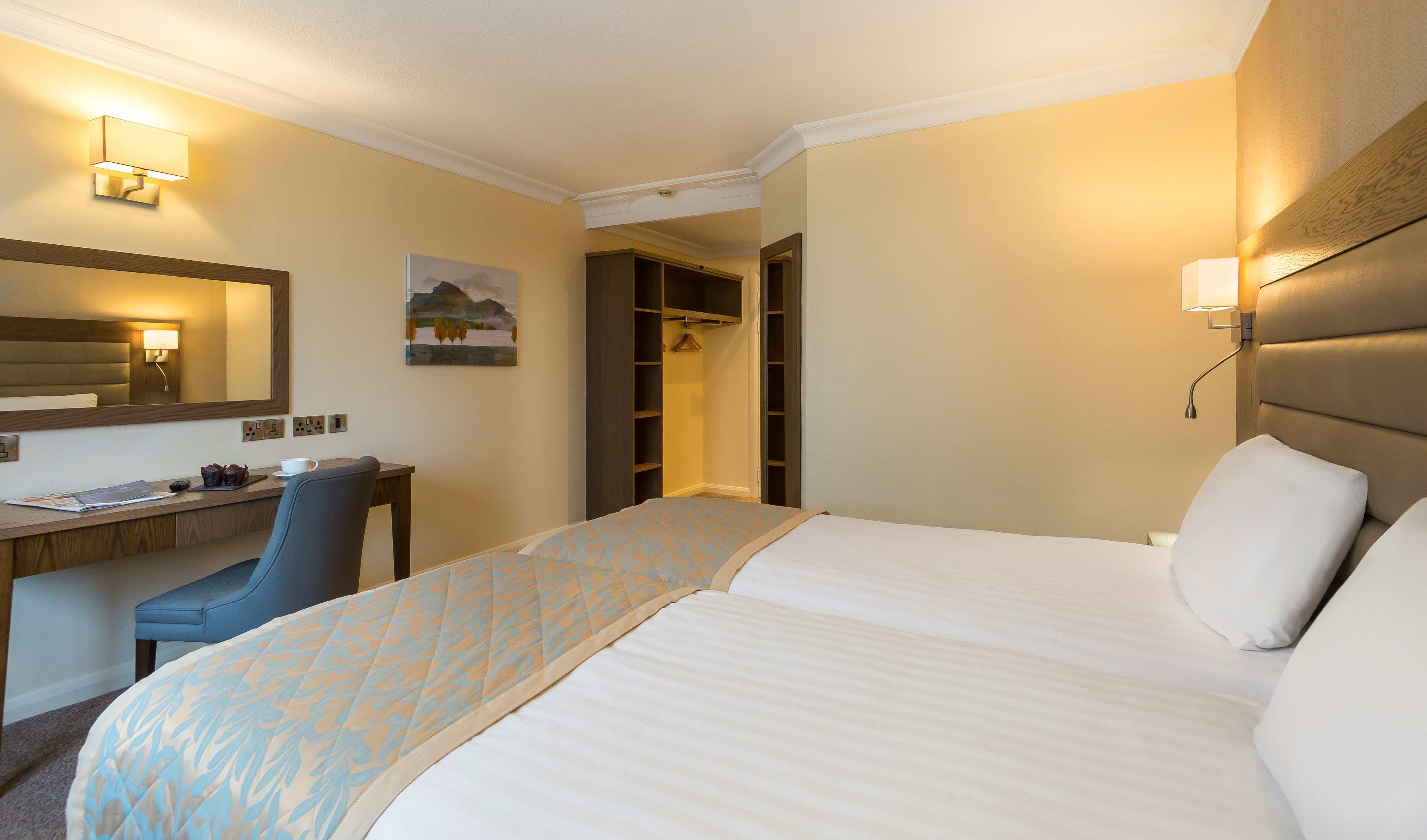 Una habitación de hotel con cama, escritorio, silla, armario y un cuadro en la pared.
