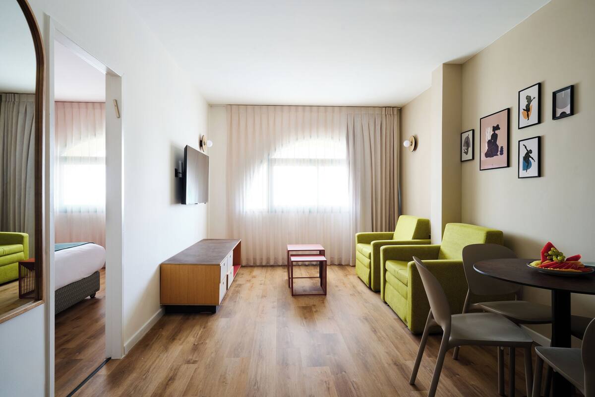 A modern hotel room with a wooden floor, a green sofa, a small table, and a TV mounted on the wall.