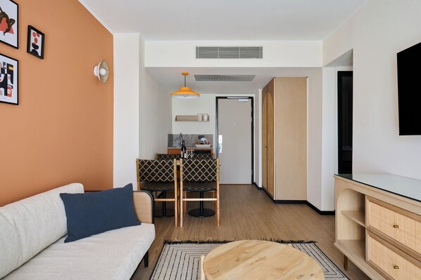 A modern living room with a sofa, a dining area with chairs, a kitchen in the background, and a TV mounted on the wall.