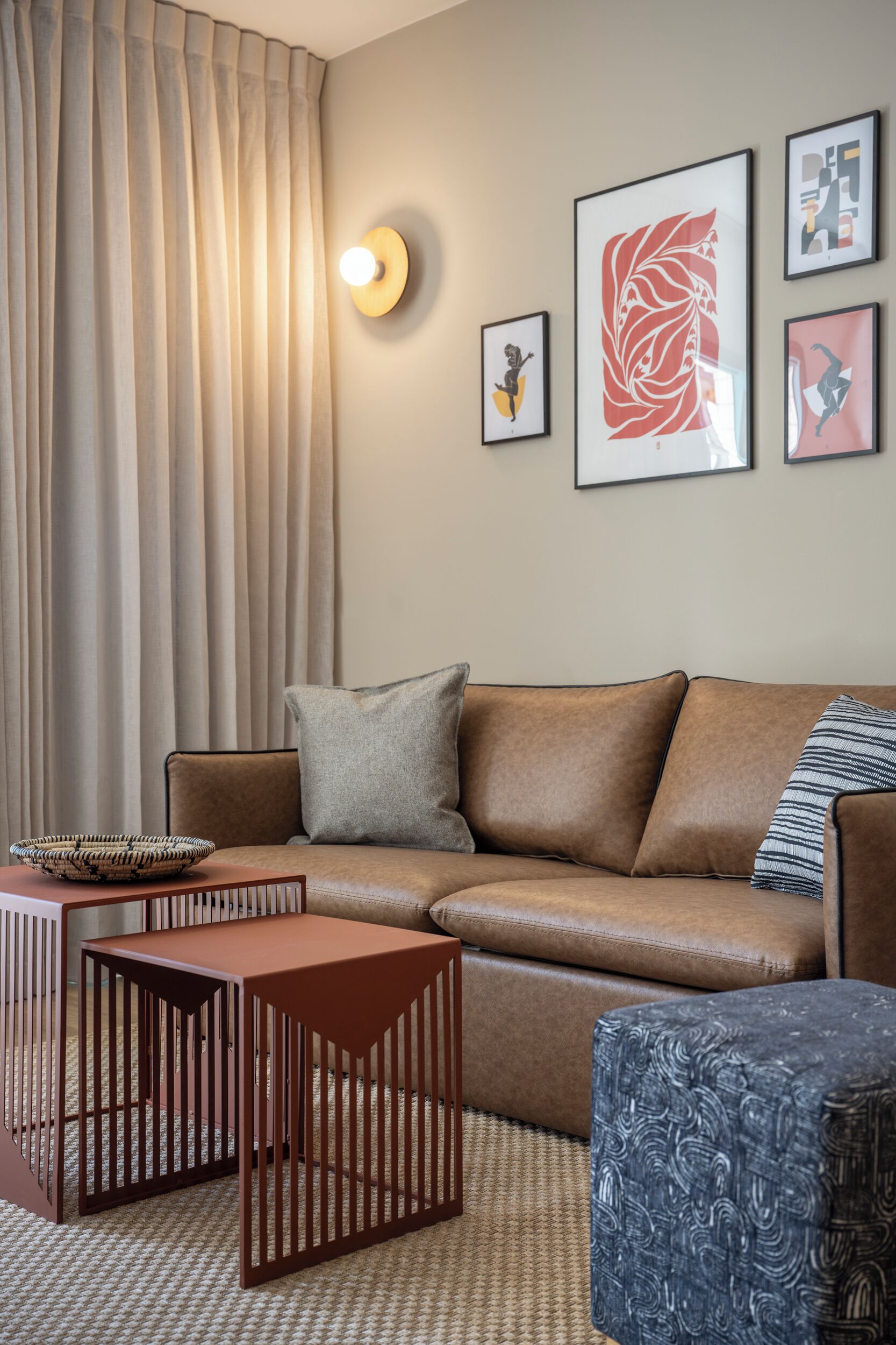 A modern living room with a brown leather sofa, a wooden coffee table, a patterned ottoman, and a woven rug. There are framed artworks on the wall and a light fixture above the sofa.