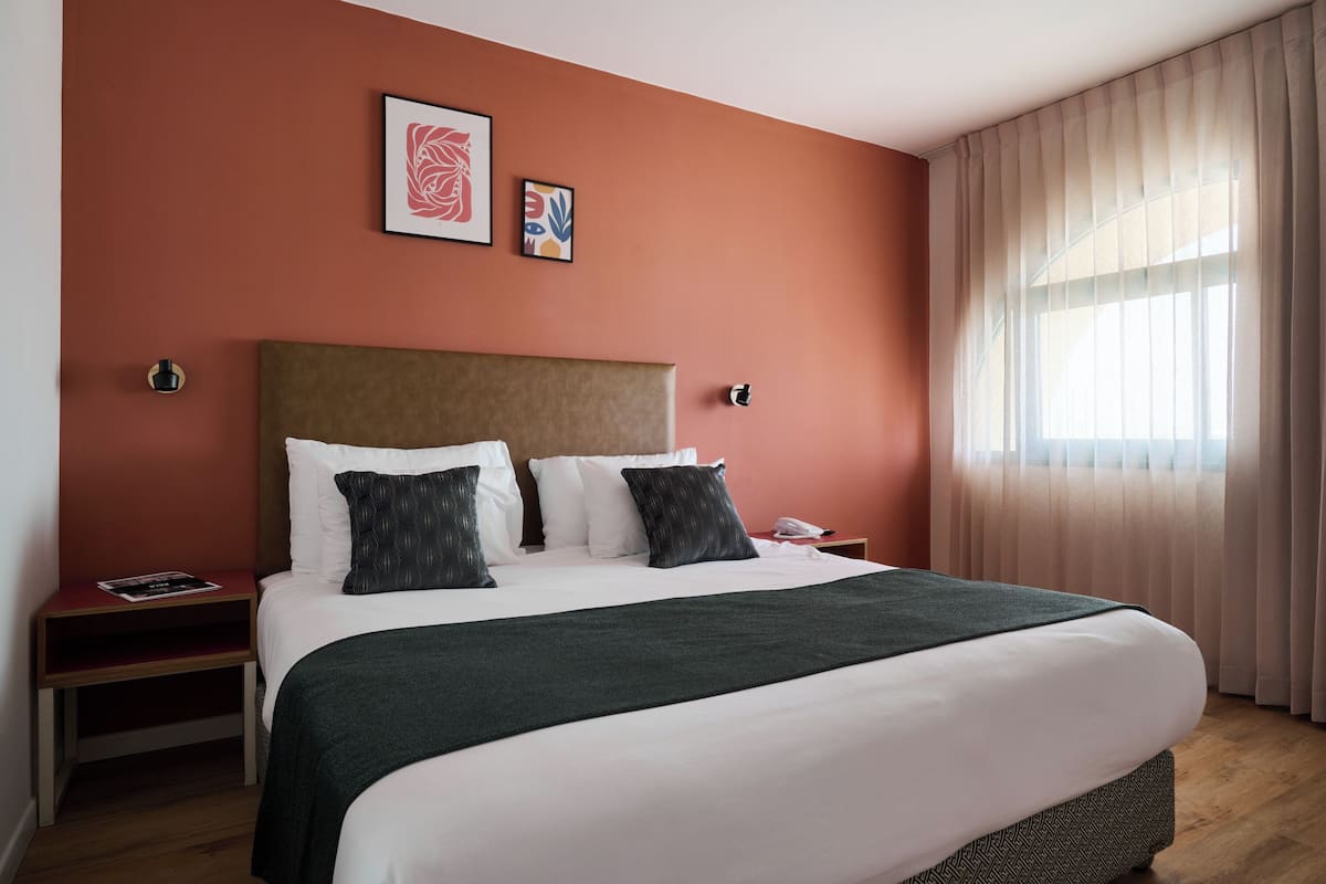 A hotel room with a large bed, a headboard, a bedside table, a nightstand, and a window with curtains.